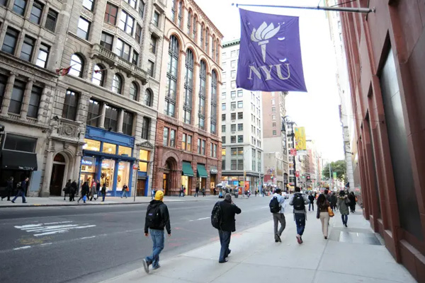 People walking down the sidewalk on the streets of New York University. A purple NYU flag draped from a building hangs high over the sidewalk. Photo credit Christopher Sadowski